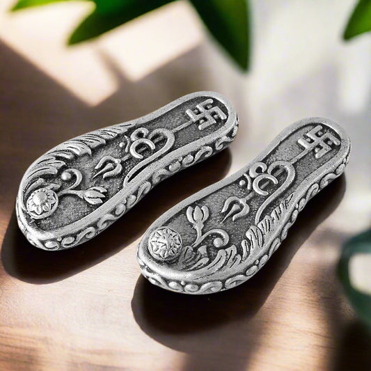 Two silver paduka with intricate designs on a white background