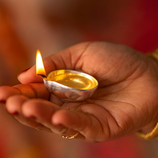 Hand holding a small silver diya with a lit wick inside against a blurred background